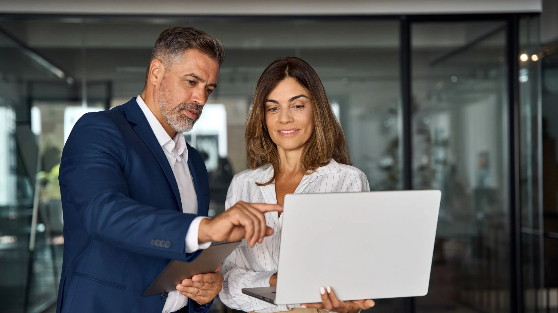 Two business professionals reviewing data on a laptop and tablet in a modern office, discussing work strategy and collaboration.