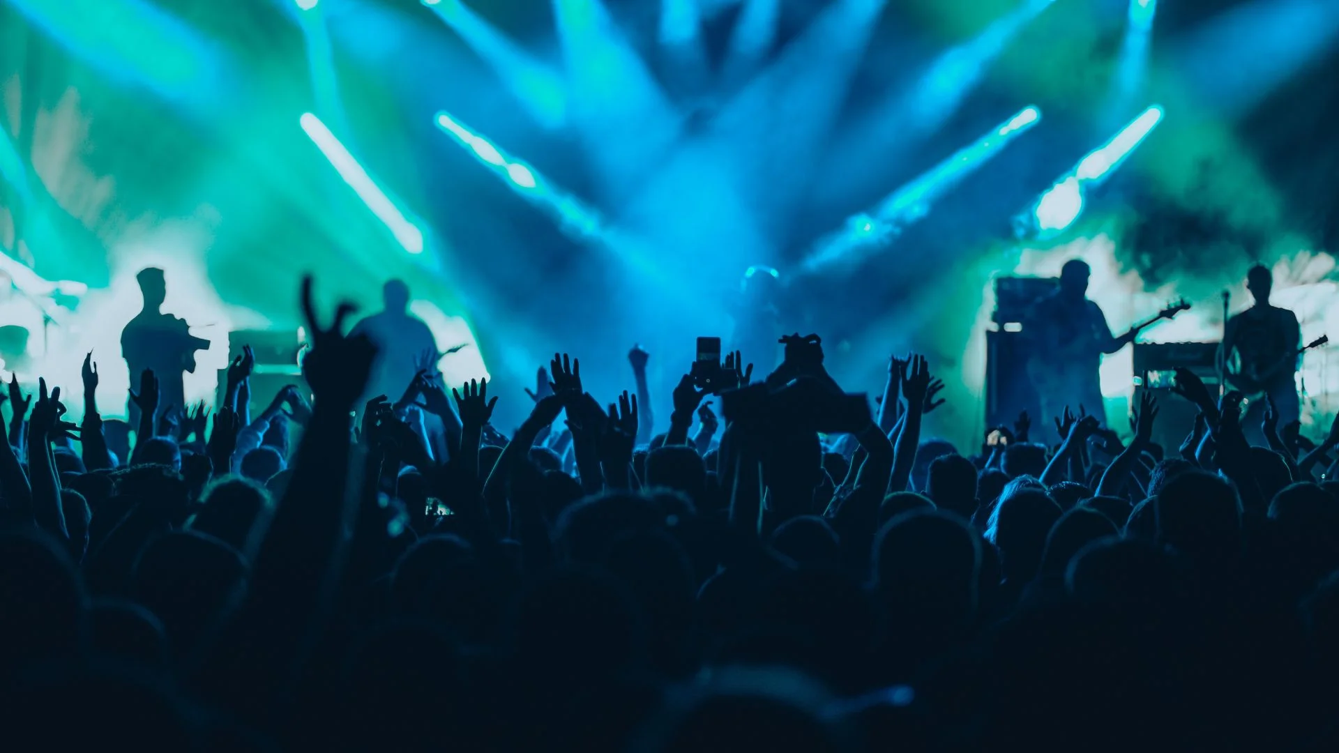 Crowd at live music concert with raised hands and band performing on stage under blue and green stage lights