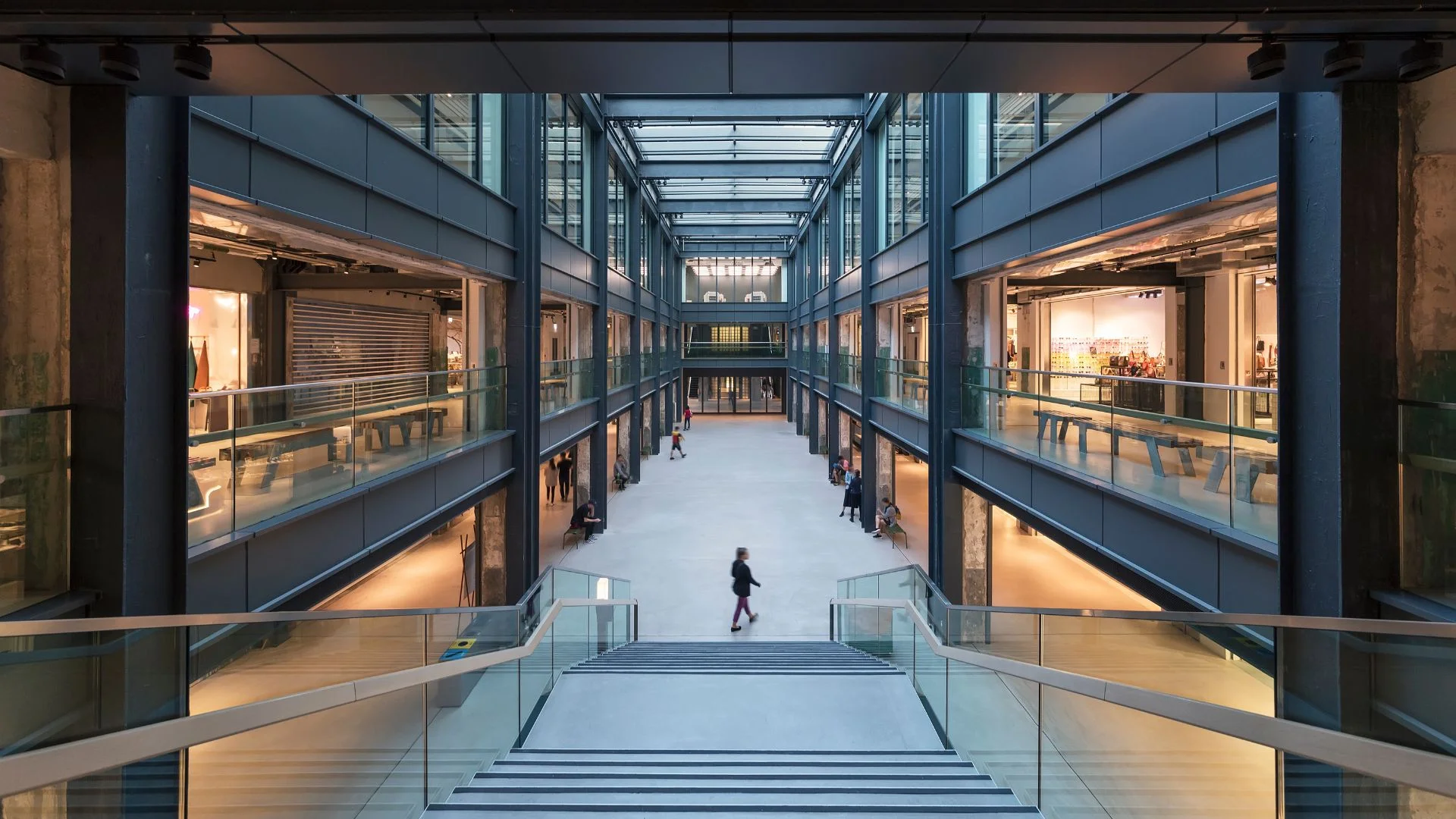Modern industrial-style shopping mall interior with glass ceiling, multiple levels, and people walking through central atrium