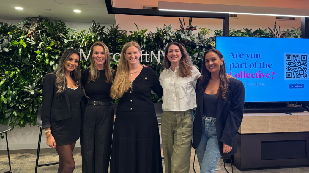 Group of five women posing and smiling in front of a green plant wall and digital screen at a professional event.