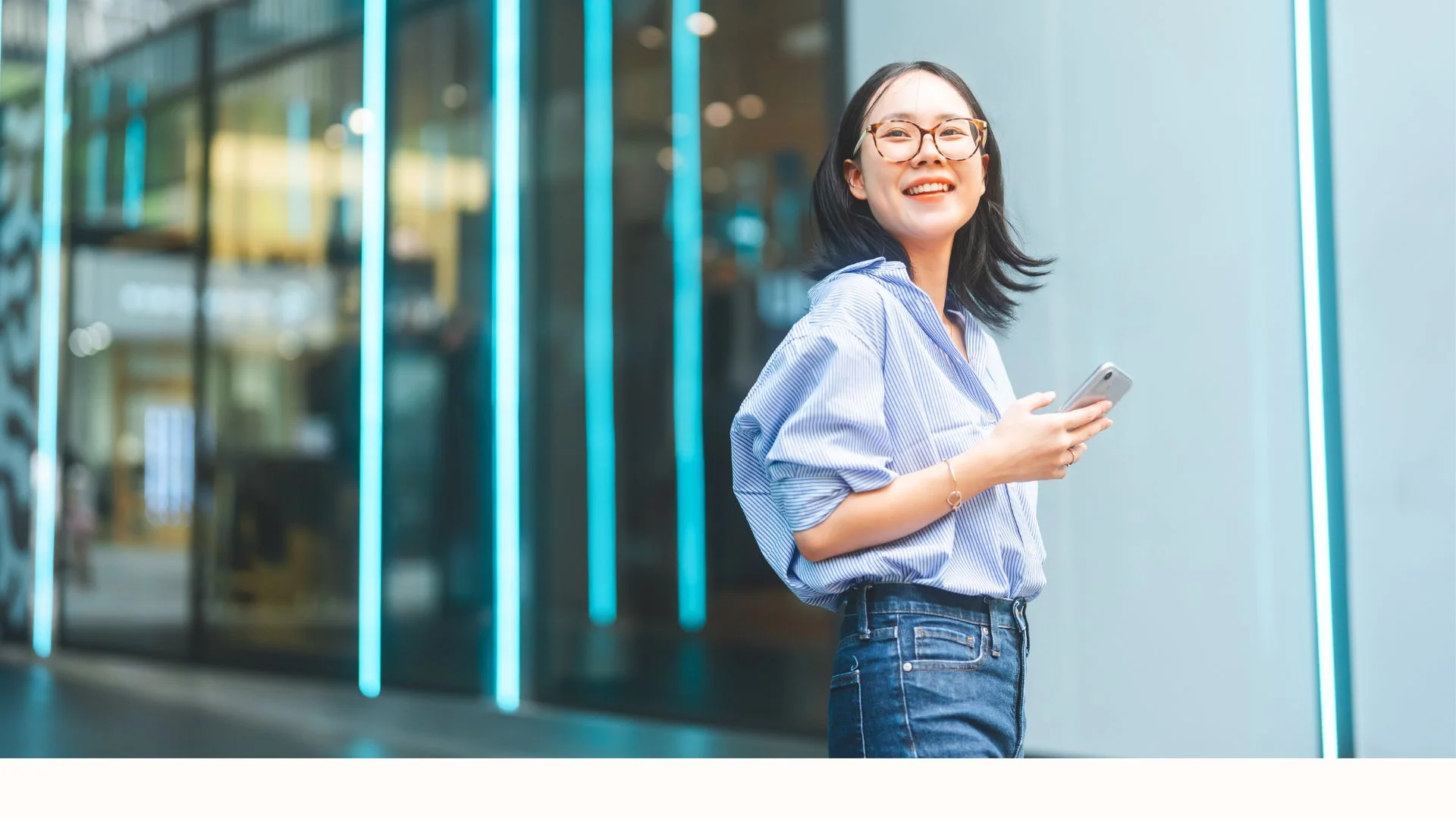 Smiling woman in glasses holding a smartphone, standing outside a modern building with blue vertical lights.