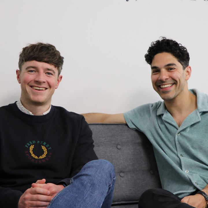 Two men sitting closely on a gray couch, smiling at the camera. The man on the left has curly black hair and wears a light green shirt, while the man on the right has dark brown hair and sports a dark sweater with a Fred Perry logo. Both are ecommerce recruiters.