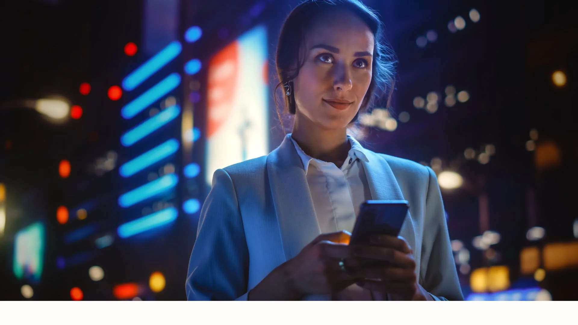 Woman in a light blue blazer using a smartphone at night in a city with bright neon lights.