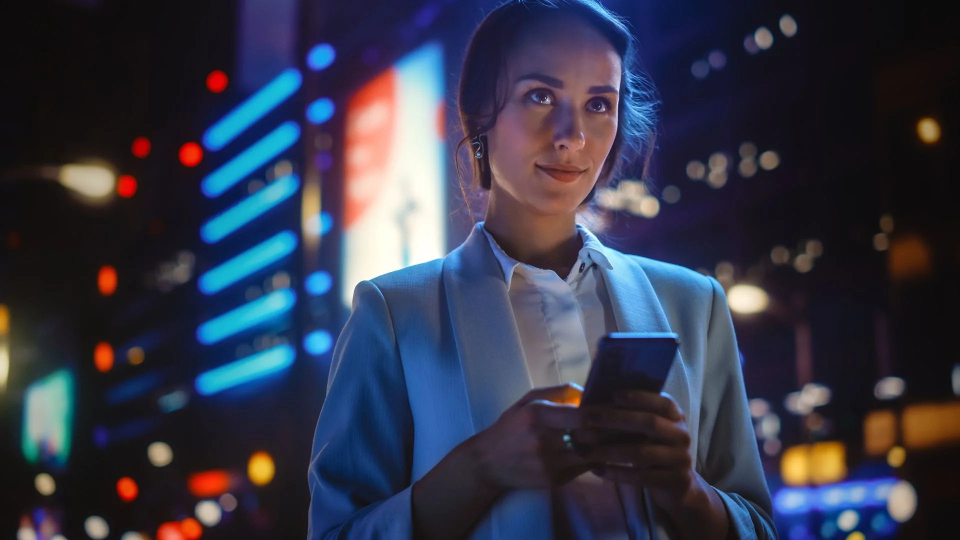 Woman in a light blue blazer using a smartphone at night in a city with bright neon lights.