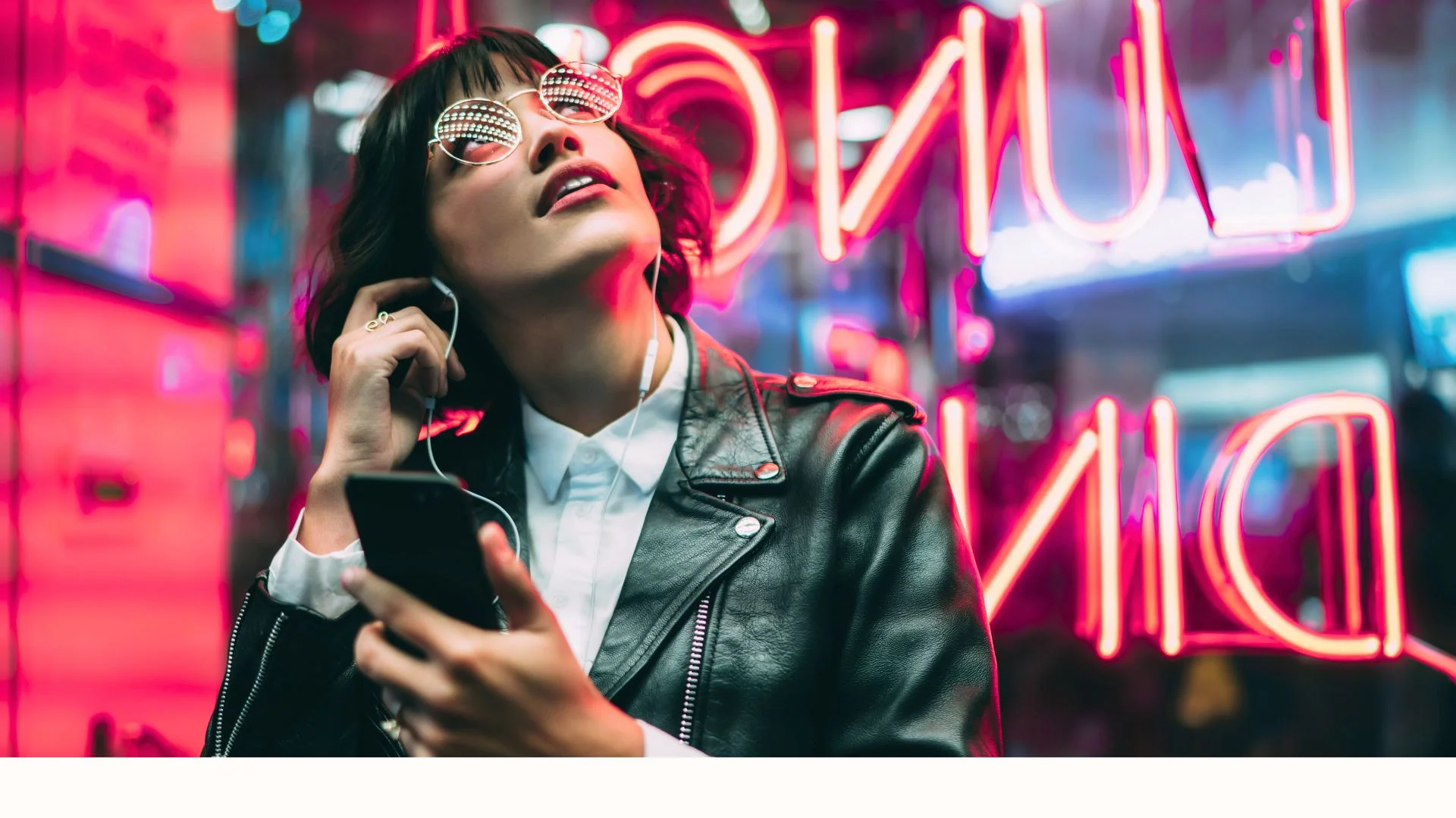 Woman in a leather jacket holding a smartphone and wearing earphones, standing in front of bright neon lights at night.