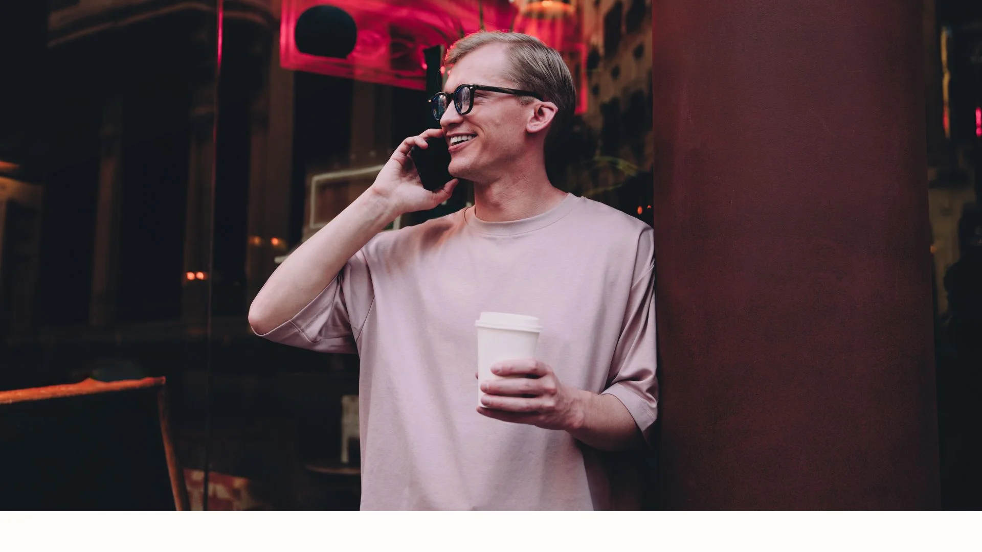 Smiling man in glasses holding a coffee cup and talking on the phone while leaning against a column in an urban setting.
