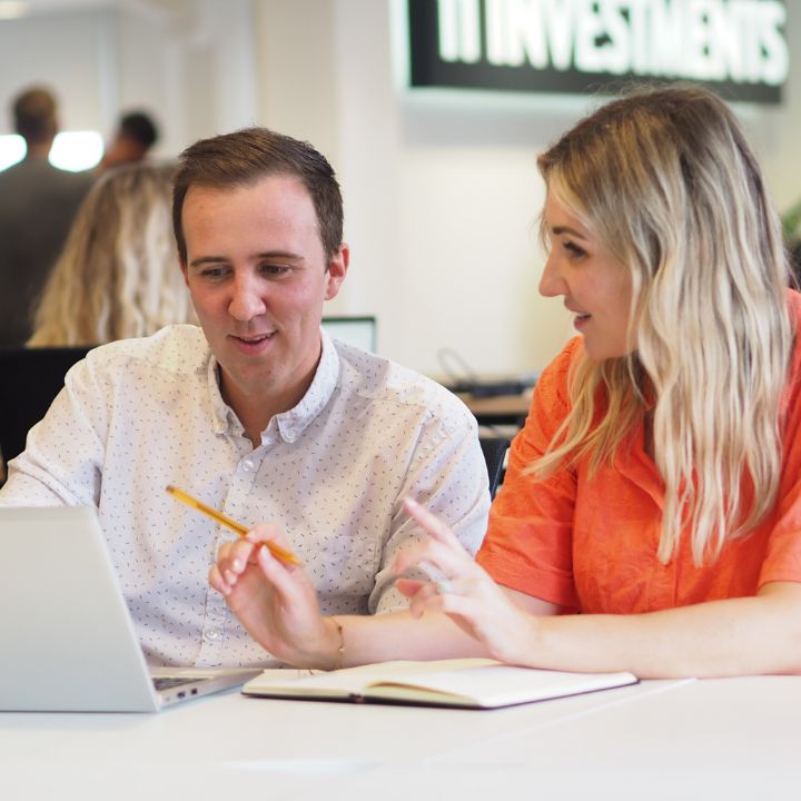 A man and a woman are seated at a white table, collaborating on a project with a laptop in a busy office environment. The man, wearing a white shirt with a subtle print, listens intently to the woman, who is dressed in a bright coral blouse and holding a pencil, as she explains a point. In the background, there's other office workers, creating a dynamic professional atmosphere.