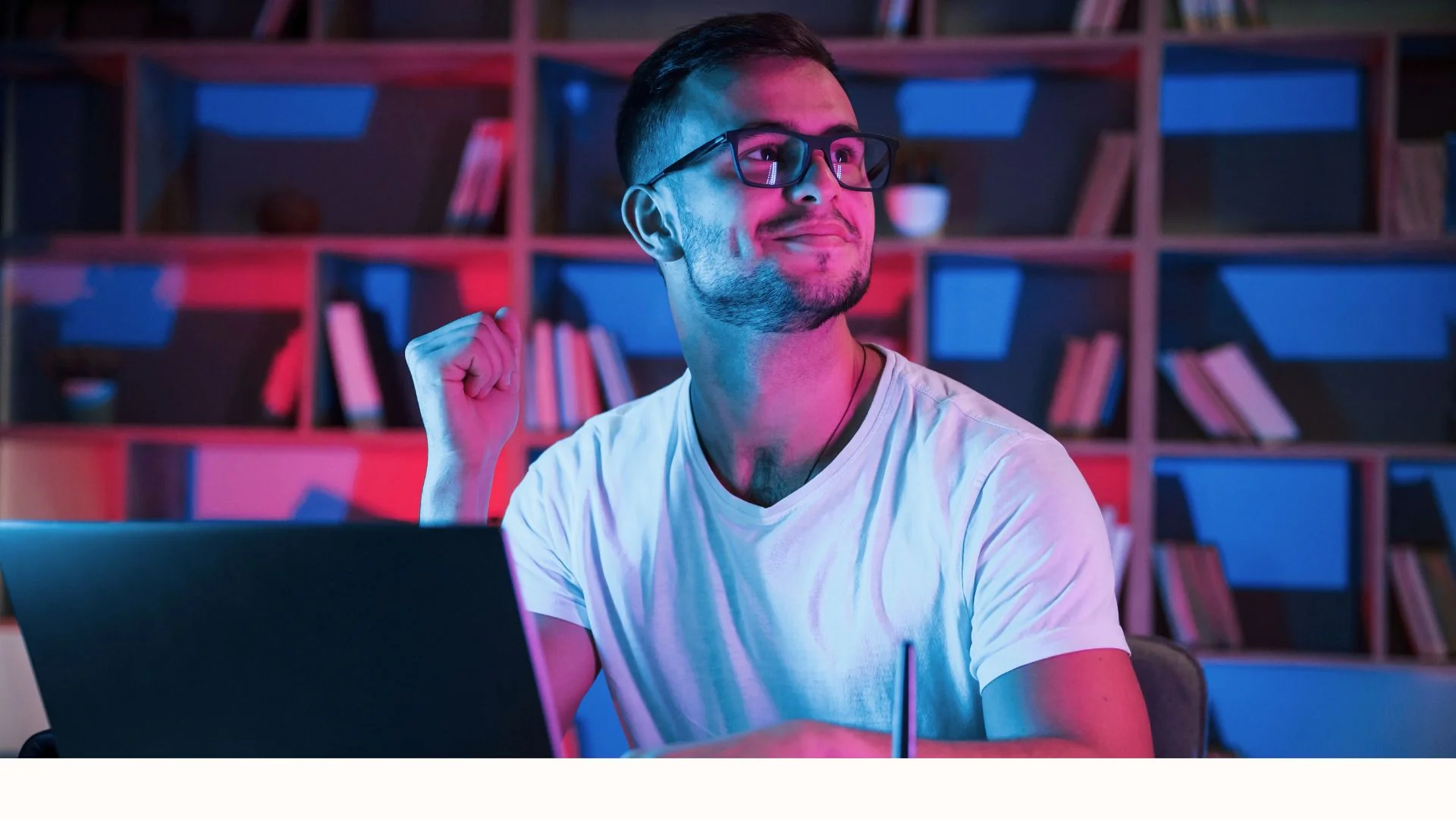 Smiling man wearing glasses sitting at a desk with a laptop, in a room with colorful lighting and bookshelves in the background.