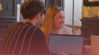 On the left of the image there is the back of a male recruiter's head as he works on a laptop. To his immediate right there is a female recruitment consultant smiling.