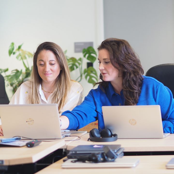 Two women are sitting next to each other at a modern office desk, each working on a laptop. The woman on the left, wearing a light yellow shirt, is smiling while looking at her screen. The woman on the right, dressed in a blue shirt, is pointing at something on her screen, seemingly explaining or discussing it with her colleague. They are surrounded by office accessories like headsets and other gadgets, suggesting a collaborative and tech-oriented environment.