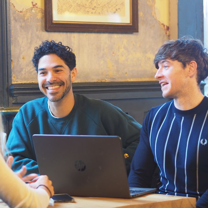 Two men sitting at a table with a laptop between then. They're smiling and talking with colleagues.