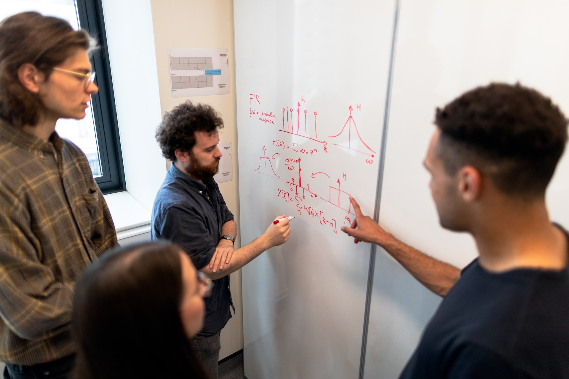 A photograph of colleagues meeting to discuss data in front of a whiteboard displaying graphs and mathematical formulae written in red marker pen.